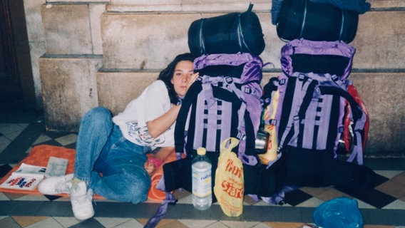 Eine junge Frau sitzt neben zwei großen Rucksäcken vor einem Bahnhof. © NDR/Pier 53 Filmproduktion 