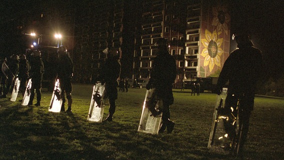 Polizisten in schwerer Ausrüstung und mit Schilden schirmen das inzwischen geräumte und teilweise abgebrannte Asylbewerberheim in Rostock-Lichtenhagen in der Nacht zum 27. August 1992 ab. Polizisten in schwerer Ausrüstung und mit Schilden schirmen das inzwischen geräumte und teilweise abgebrannte Asylbewerberheim in Rostock-Lichtenhagen in der Nacht zum 27. August 1992 ab. © picture alliance / ZB Foto: Jens Kalaene