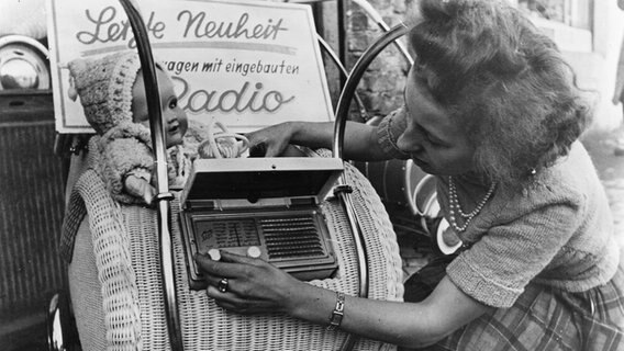Eine Frau dreht 1950 an einem Radio, das in einen Kinderwagen eingebaut ist. Das Kofferradio "Metz Baby" wurde als "letzte Neuheut" beworben. © picture alliance / akg-images 