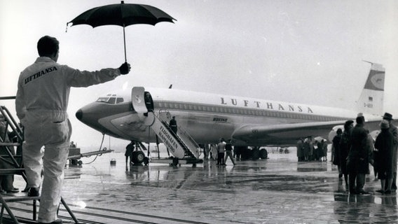 Ein Lufthansa-Mitarbeiter hält einen Regenschirm, während eine Boeing 707 der Lufthansa auf dem Rollfeld des Hamburger Flughafens steht. © imago images 