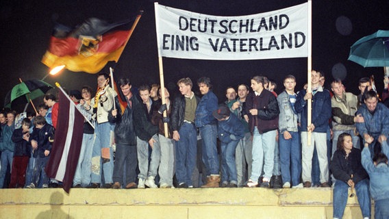 Mit der Deutschlandfahne und einem Transparent "Deutschland Einig Vaterland" stehen zahlreiche Berliner am 22. Dezember 1989 auf der Berliner Mauer am Brandenburger Tor. © dpa Foto: dpa