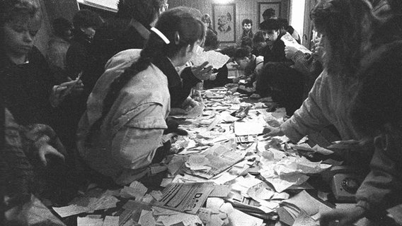 Menschen stehen am 4. Dezember 1989 in der Stasi-Zentrale in Erfurt (Thüringen) an einem Tisch voll mit durchwühlten Unterlagen der Behörde. © picture alliance / dpa | Gesellschaft für Zeitgeschichte 