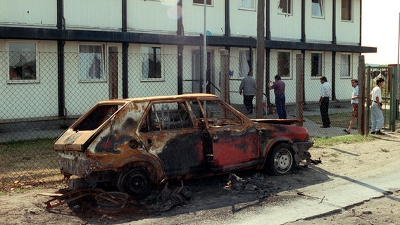 Ein ausgebranntes Auto steht 1991 vor dem Asybewerberheim in Ueckermünde. © picture-alliance / ZB Foto: Kalaene Jens