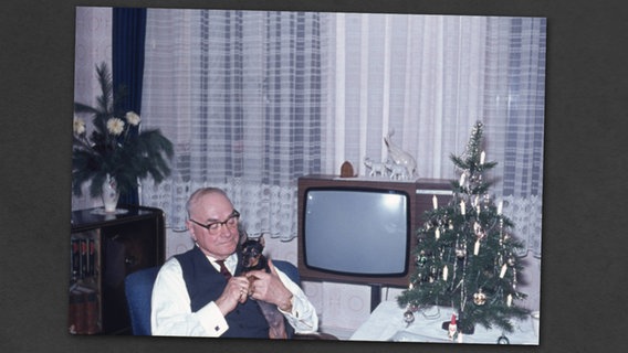 Ein älterer Mann sitzt mit seinem Hund auf dem Arm neben einem Weihnachtsbaum. © imago stock&people 