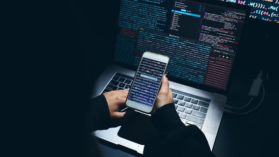 Hände halten ein Smartphone vor einem Laptop. Auf den Bildschirmen befinden sich abstrakte Codes. © photocase.de Foto: przemekklos