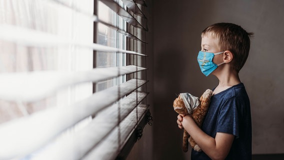 Ein kleiner Junge mit Maske und einem Teddy im Arm schaut aus einem Fenster. © imago images/Cavan Images 