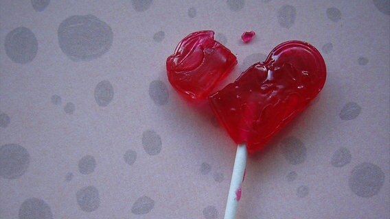 Ein roter Herzlolly mit weißem Stiel liegt zerbrochen auf einer grauen Unterlage. © knallgrün / photocase.de 
