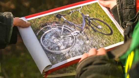 Zwei Personen betrachten ein Bild, auf dem der Akku eines Rades gebrannt hat. © NDR Foto: Screenshot