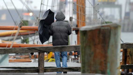 Ein Mann mit Kapuze und Regenschirm geht am Hafen spazieren. Ein Mann mit Kapuze und Regenschirm geht am Hafen spazieren. © dpa Picture Alliance Foto: Carsten Rehder
