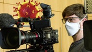 Ein Kameramann mit Mundschutz, im HIntergrund eine Grafik des Coronvirus. © NDR 