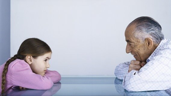Ein junges Mädchen mit Zöpfen und ein älterer Mann sitzen sich auf die Arme nach vorne gestützt an einem Tisch gegenüber. © Colourbox Foto: -