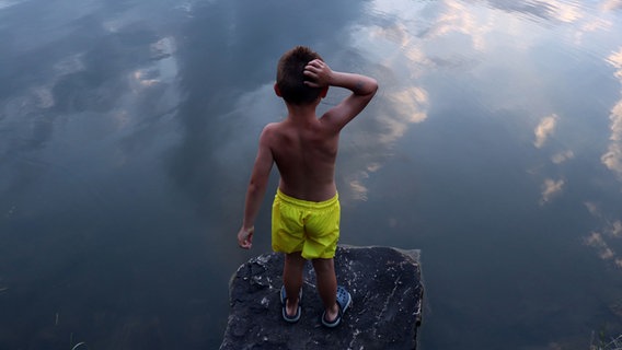 Ein Junge in gelber Badehose steht auf einem Felsen am Wasser und kratzt sich am Hinterkopf. © photocase/fotokomplizin Foto: fotokomplizin