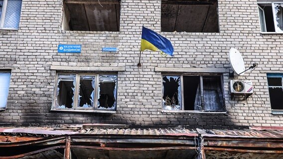 An einer zerstörten Hauswand hängt eine ukrainische Flagge. © picture alliance_AA Foto: Yevhen Titov