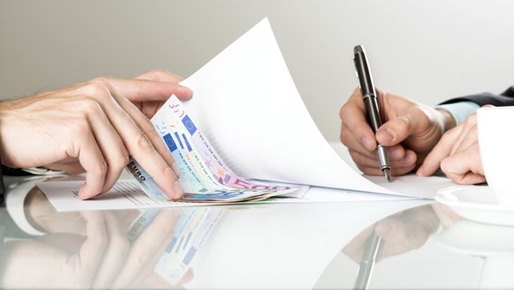 Ein Mann schiebt Geldscheine unter ein, auf einem Schreibtisch liegendes Papier, welches zeitgleich von einer anderen Person unterzeichnet wird. © panthermedia Foto: halfpoint