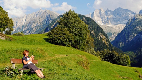 Frau liest ein Buch auf einer Bank - im Hintergrund eine Bergkulisse © picture alliance / imageBROKER | Oliver Gerhard 