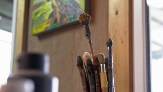 Ein Gefäß mit Pinseln steht in einem Atelier, im Hintergrund hängt ein Bild an der Wand. © NDR 