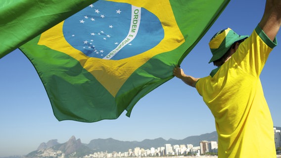 Ein Mann steht vor der Skyline von ipanema mit eine Brasilien Fahne in den Händen. ©  lazyllama/fotolia Foto:  lazyllama