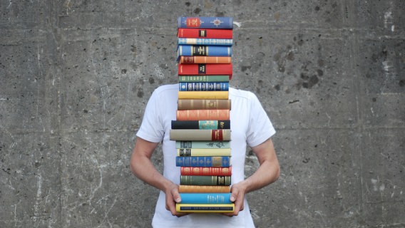Ein junger Mann trägt einen hohen Stapel Bücher der seinen Kopf verdeckt. © photocase.de Foto: luxuz