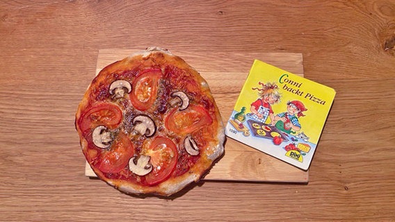 Ein Buch aus der Reihe "Connie" liegt neben einer ganzen runden Pizza © NDR Foto: Katharina Mahrenholtz