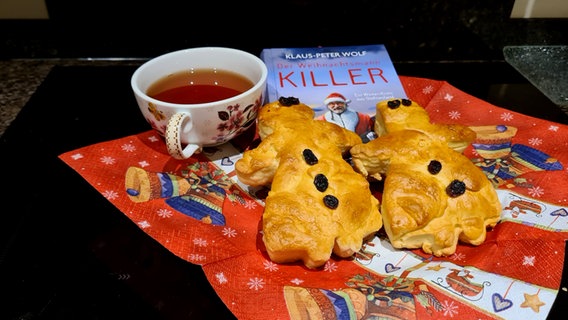 Eine Tasse Tee auf einer Weihnachtsserviette, darauf liegen zwei Gebäckstücke Stutenmänner und ein Buch © NDR Foto: Jan Ehlert