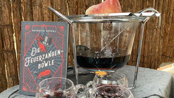 Das Buch von Heinrich Spoerl "Die Feuerzangenbowle" steht auf einem Tisch neben einer Feuerzangenbowle im Freien © NDR 