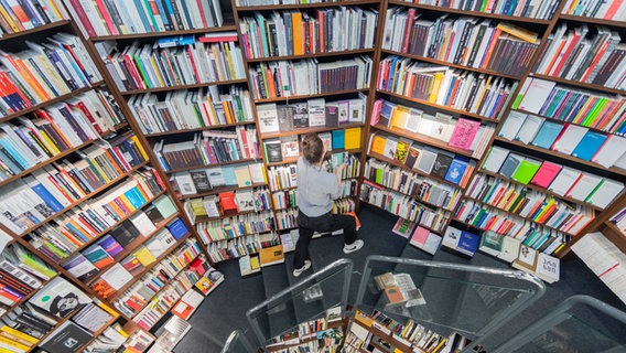 Viele Bücher in Regalen in einer Buchhandlung, eine junge Frau steht vor einem der Borde und zieht Bücher heraus © Rolf Vennenbernd/dpa +++ dpa-Bildfunk +++ Foto: Rolf Vennenbernd