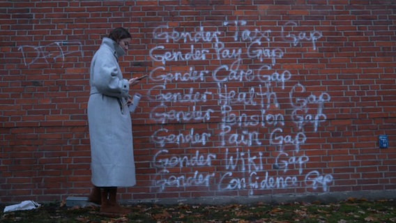 Eine Frau steht vor einer Backsteinwand, auf die Wörter gesprayt wurden © Screenshot NDR 