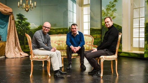 Drei Männer sitzen auf Stühlen auf einer Bühne. © Theater Lübeck Foto: Jan Philip Welchering