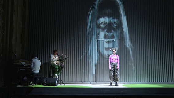 Eine Frau steht auf einer Bühne, im Hintergrund sitzen zwei Musiker © Screenshot NDR 