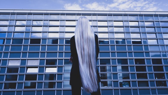 Eine Frau mit hellen langen Haaren, die bis zu den Oberschenkeln reichen, steht mit dem Rücken zur Kamera vor einem modernen Gebäude - Themenfoto zur Aufführung "Noch wach?" am Thalia Theater © Krafft Angerer 