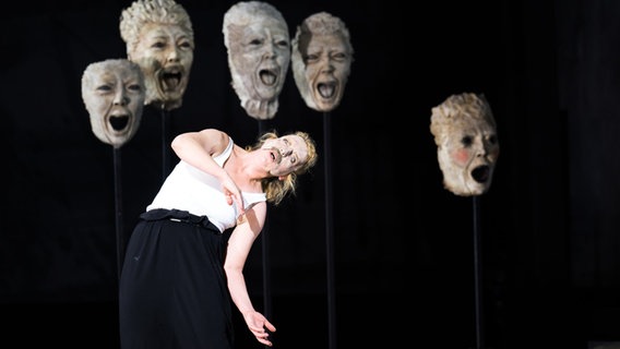 Eine Frau mit Maske auf einer Bühne - im Hintergrund große Masken. Eine Frau mit Maske auf einer Bühne - im Hintergrund große Masken. © Monika Rittershaus Foto: Monika Rittershaus