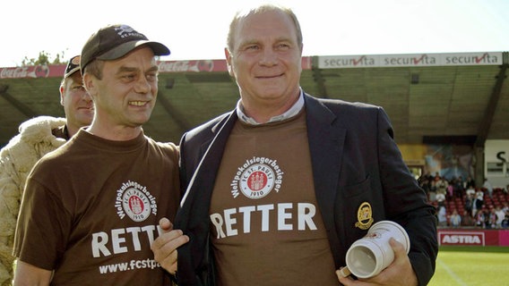 Bayern-München-Manager Uli Hoeneß (re.) präsentiert sich am 12. Juli 2003 neben St.-Pauli-Präsident Corny Littmann beim Benefizspiel zur Lizenzsicherung des FC St. Paulis im Retter-T-Shirt. © IMAGO / HochZwei 