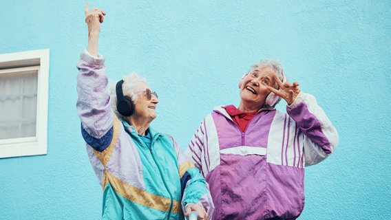 Zweite ältere Frauen in Trainingsanzug tanzen und hören Musik. © IMAGO / Zoonar 