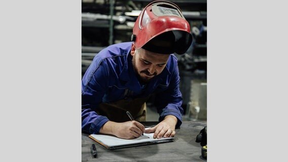 Ein Mann mit einem Schweißerhelm auf dem Kopf trägt einen Blaumann. Er beugt sich über einen Arbeitstisch, auf dem ein Block liegt. Mit einem Stift in der Hand schreibt er darauf. © Addictive Stock / photocase.de 