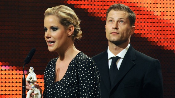 Eine blonde Frau mit gepunktetem Oberteil schaut zur linken Seite und spricht in ein Mikrofon. In der Hand hält sie eine Trophäe. Rechts hinter ihr steht ein Mann im schwarzen Anzug, den Blick nach oben gerichtet. © picture alliance/dpa | Tobias Hase Foto: Tobias Hase