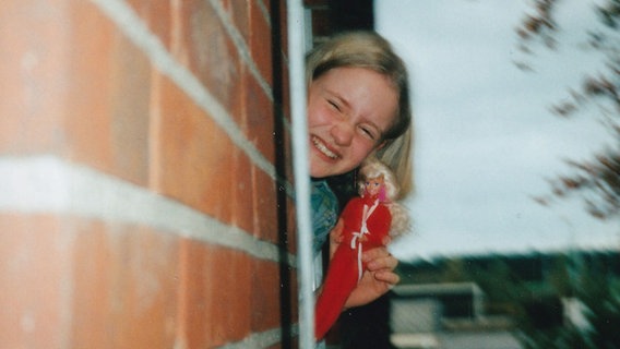 Ein junges Mädchen schaut aus einem Fenster und hält dabei eine Barbie nach oben. © Juliane Bergmann / NDR 