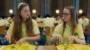 Zwei Schülerinnen in grüngelben Uniformen halten Besteck über ihren ihren Essenstabletts, die nur ein Stück Kartoffel enthalten (Szene aus "Club Zero" von Jessica Hausner) © Neue Visionen Filmverleih 