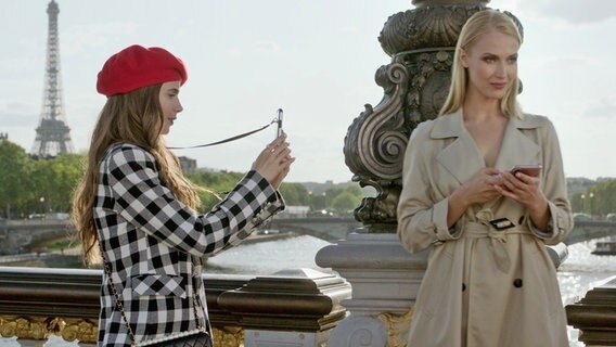 Lilly Collins (links) als Emily macht ein Foto auf einer Brücke über der Seine in Paris, im Hintergrund der Eiffelturm - Szene aus "Emily in Paris", die Serie ©  COURTESY OF NETFLIX © 2020 