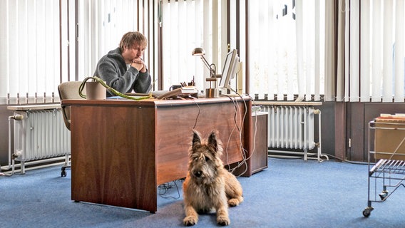 Sörensen (Bjarne Mädel) sitzt am Schreibtisch. Davor liegt Hund Cord. © Claussen+Putz Filmproduktion GmbH Foto: Michael Ihle