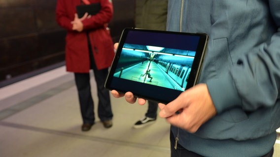 Arne Krasting hält ein Tablet - darauf ist eine Filmszene von "Cortex" in der U-Bahn Hafencity zu erkennen -  mit Jannis Niewöhner auf einer Bank, daneben eine U-Bahn © NDR Foto: Patricia Batlle