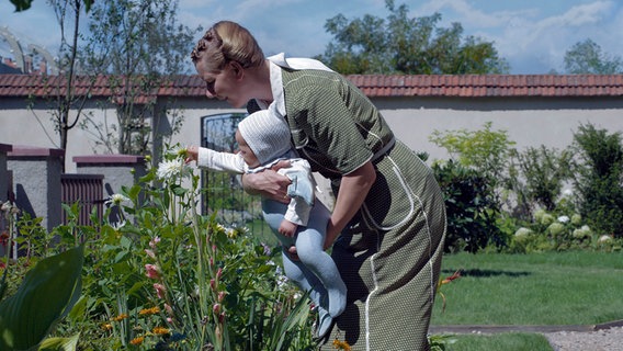 Eine Frau hält ein Baby vor sich und beugt sich über ein Blumenbeet. © picture alliance Foto: ASSOCIATED PRESS | Uncredited