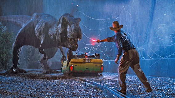 Sam Neill im Abenteuerfilm "Jurassic Park" von 1993 von Steven Spielberg mit einem umgeworfenen Auto und einem T-Rex © picture alliance / Everett Collection | ©Universal/Courtesy Everett Collection 