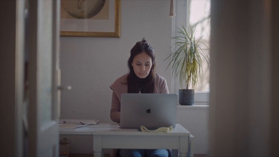 Eine Frau sitzt an einem Tisch und schaut auf einen Laptop. © NDR Screenshot 