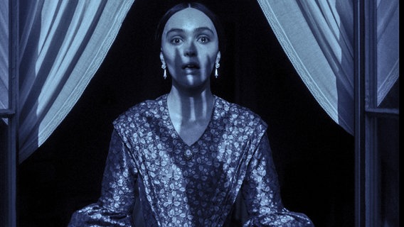 Eine Frau guckt voller Angst in einem Fenster, alles ist dunkel, über ihrem Gesicht liegt der Schatten einer Kralle von einem Ungeheuer - Szene aus "Nosferatu" von Dave Eggers © 2023 FOCUS FEATURES LLC. ALL RIGHTS RESERVED. 