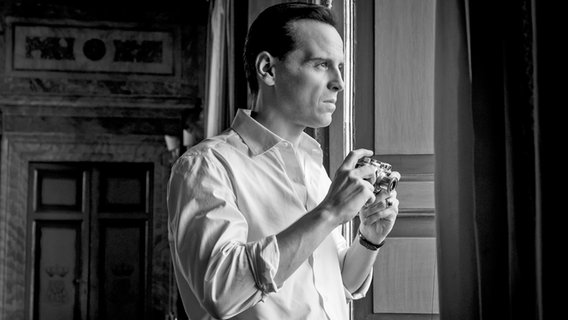 Ein Mann beobachtet hochkonzentriert aus einem Fenster etwas und hält dabei eine Kamera in der Hand - Szene aus der Szene "Ripley" mit Andrew Scott in schwarz-weiß © Philippe Antonello/NETFLIX Foto: Philippe Antonello