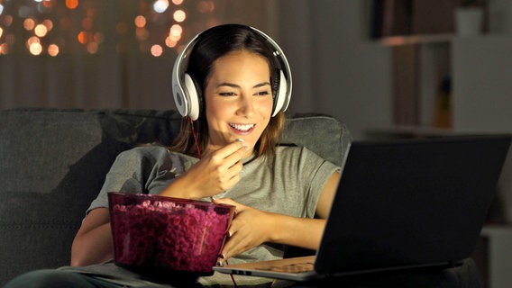 Eine Frau isst Popcorn, lächtelt und schaut auf ihrem Laptop einen Film mit Kopfhörern © IMAGO / Panthermedia Foto: Antonio Guillem