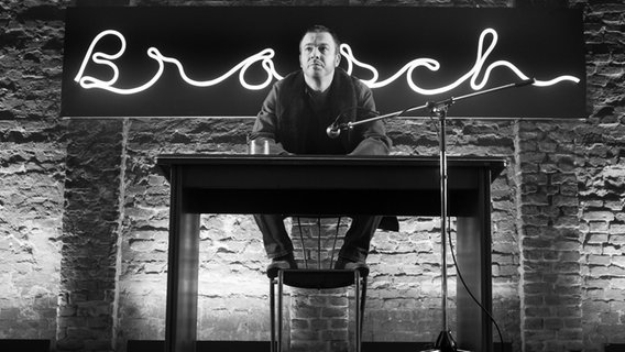 Schwarz-Weiß-Fotografie eines Mannes der auf einer Bühne an einem Tisch sitzt und nach vorne blickt. © Zeitsprung Pictures / Wild Bunch Germany Foto: Peter Hartwig
