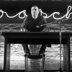 Schwarz-Weiß-Fotografie eines Mannes der auf einer Bühne an einem Tisch sitzt und nach vorne blickt. © Zeitsprung Pictures / Wild Bunch Germany Foto: Peter Hartwig