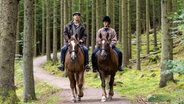 "Riding in Darkness - Der Prozess": Tommy (Jonas Karlsson) und Lotta (Malin Persson) reiten auf zwei braunen Pferden über einen Waldweg. Sie haben beide einen ernsten Gesichtsausdruck und schauen sich nicht an. © ZDF/Niklas Maupoix 