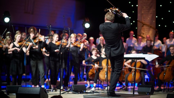 Das folkBALTICA Ensemble bei einem Konzert, im Vordergrund auf einem Pult ein Dirigent, der Geige spielt. © Folk Baltica 
